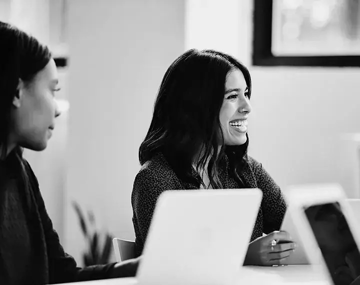 Two women engaged in work, seated at a table with laptops open in front of them, focused on their tasks.