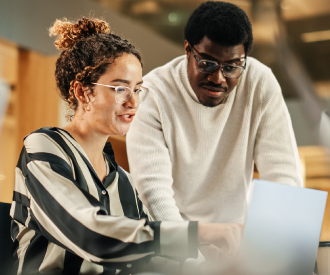 Two people work together at a desk, looking at a laptop screen. The woman is seated and typing, while the man stands next to her, both focused on the computer.