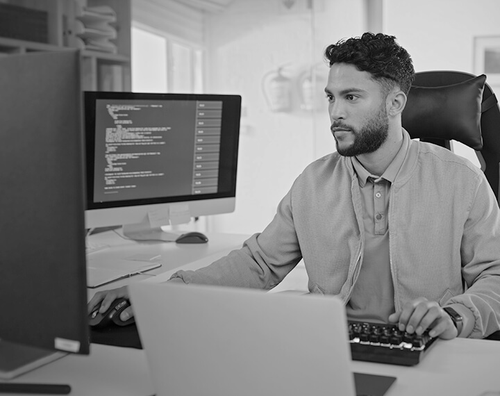 1. A man seated at a desk, working on a computer and a laptop, focused on his tasks in a professional setting.