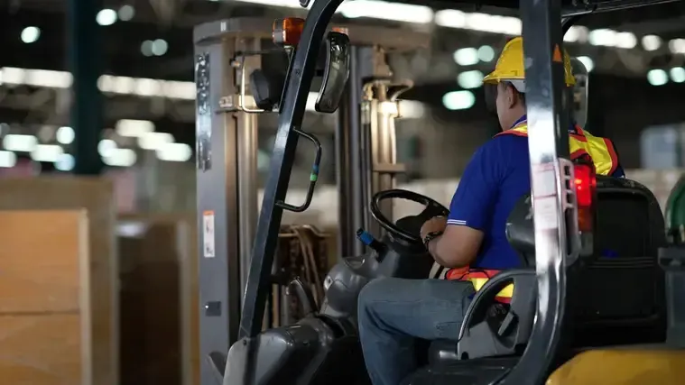 A worker operates a forklift inside a warehouse, maneuvering through stacks of goods and equipment.